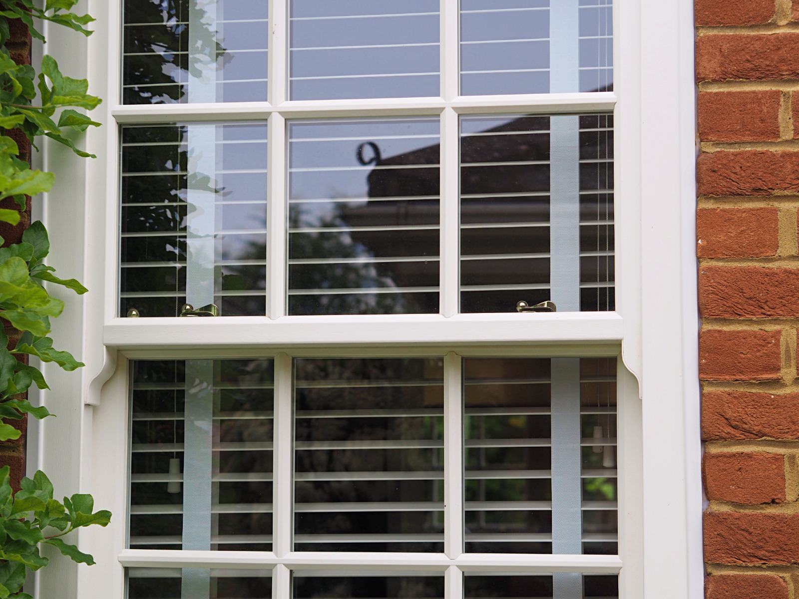 Close-up of a white uPVC sliding sash window with Georgian glazing bars and brass fitch fasteners in a red-brick wall