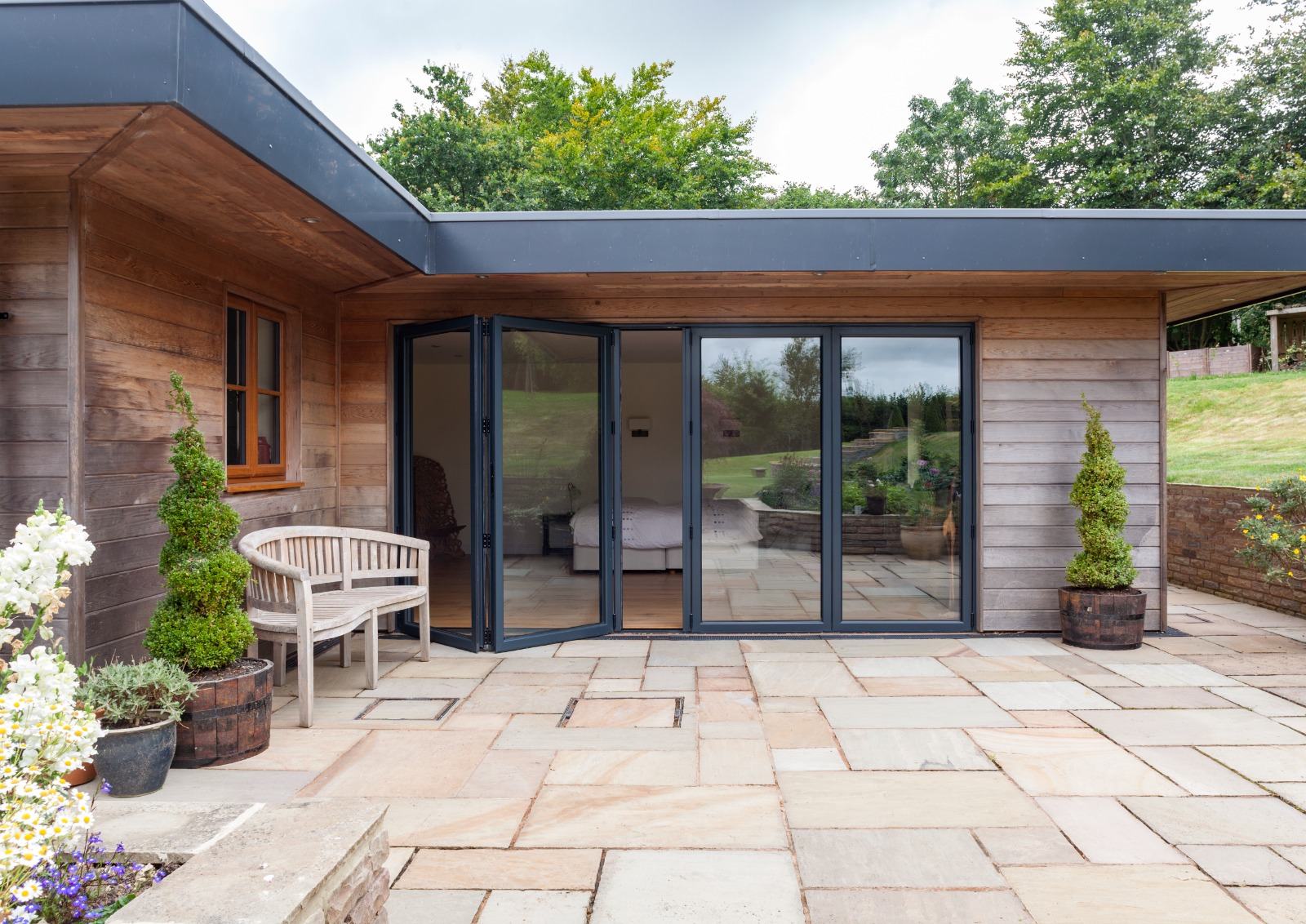Oak-clad garden room with anthracite grey aluminium bi-fold doors and matching sliding window opening onto a sandstone patio